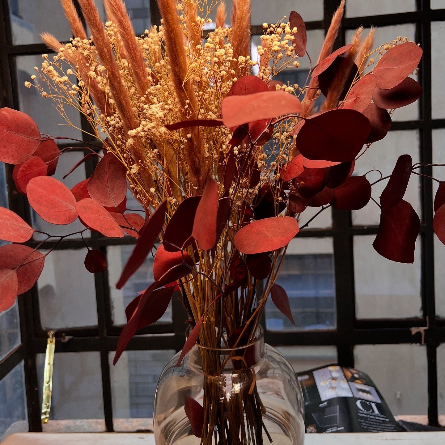 Bouquet of red and gold leaves in a clear vase against a window with black frames.