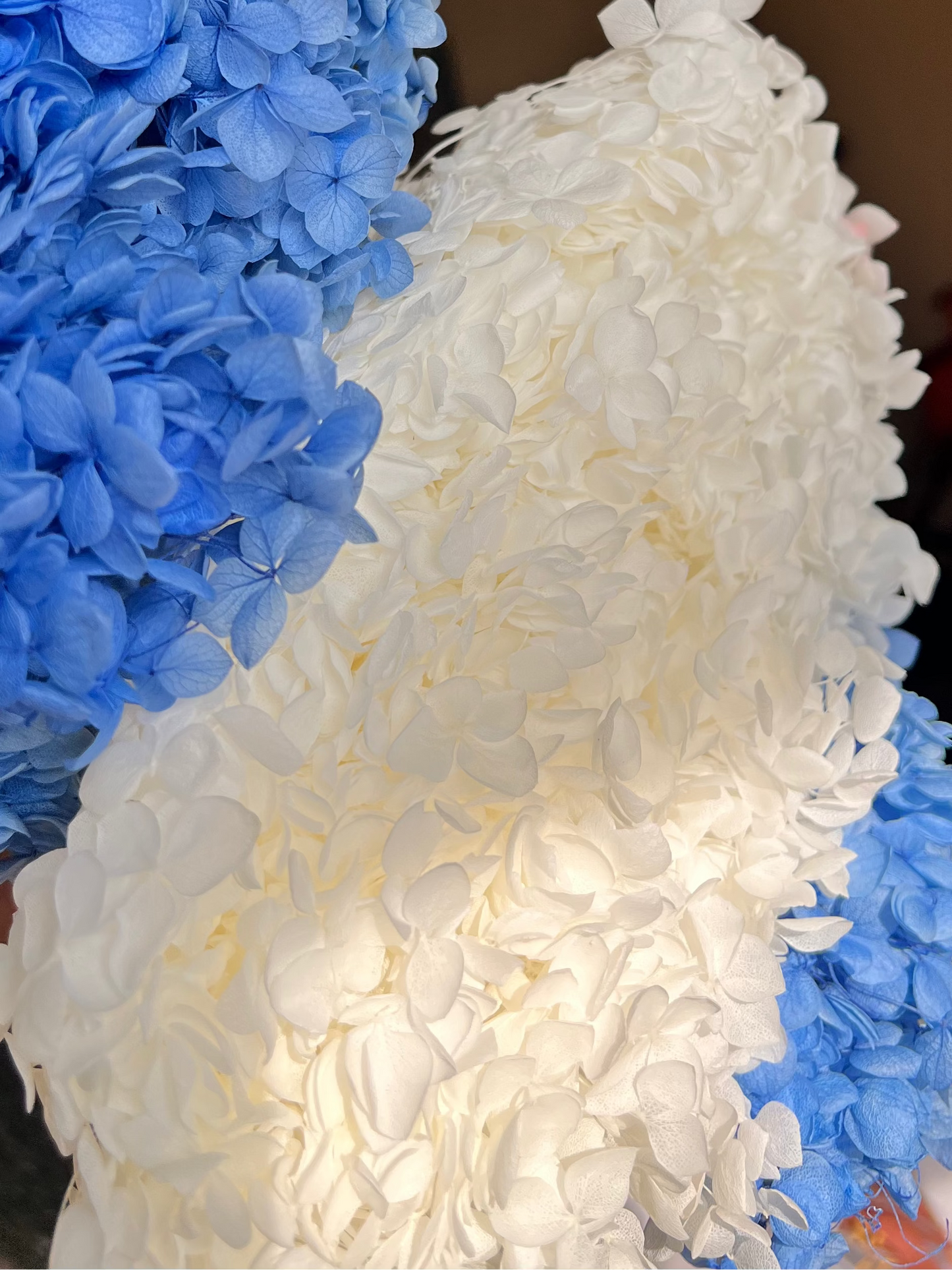 A close-up of a decorative floral arrangement featuring textured blue and white preserved hydrangea blooms from kenyan lady fleur dried flowers kenya , photographed indoors with a blurred room background.