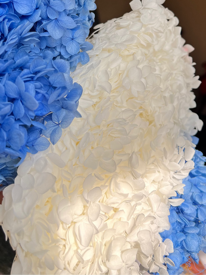 A close-up of a decorative floral arrangement featuring textured blue and white preserved hydrangea blooms from kenyan lady fleur dried flowers kenya , photographed indoors with a blurred room background.
