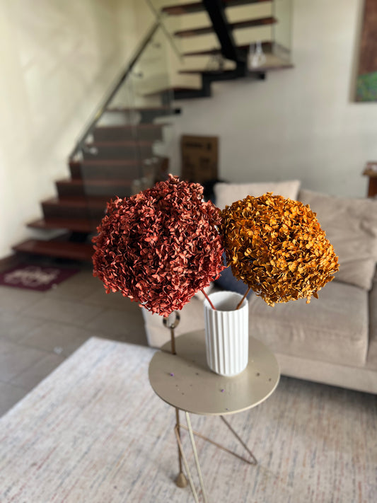 Two dried flower hydrangea bouquets in earthy colours from kenyan lady fleur dried flowers kenya in a white ribbed vase on a small round side table, set in a living room with a staircase and sofa in the background.