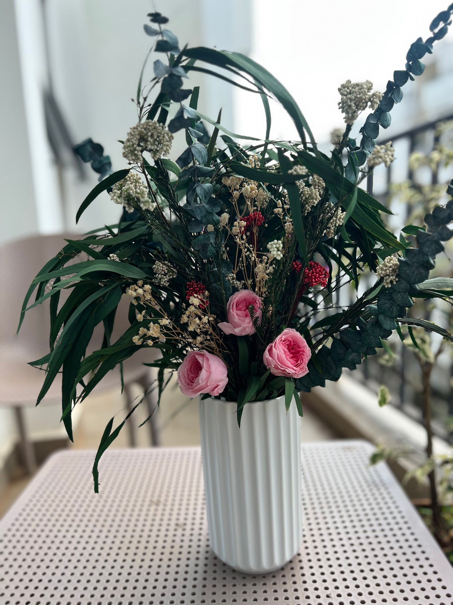 A dried floral arrangement in a white ribbed vase, featuring green eucalyptus leaves, small white clusters, pink roses, and red berries, displayed on a white perforated table from kenyan lady fleur dried flowers kenya.