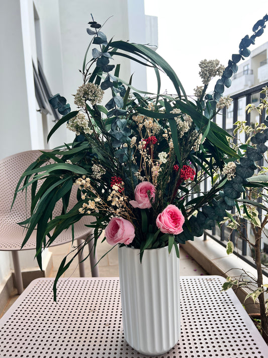A large floral arrangement in a white ribbed vase, featuring green eucalyptus, long green leaves, pink roses, and small cream and red flowers, from kenyan lady fleur dried flowers kenya displayed on a white perforated table on a balcony.
