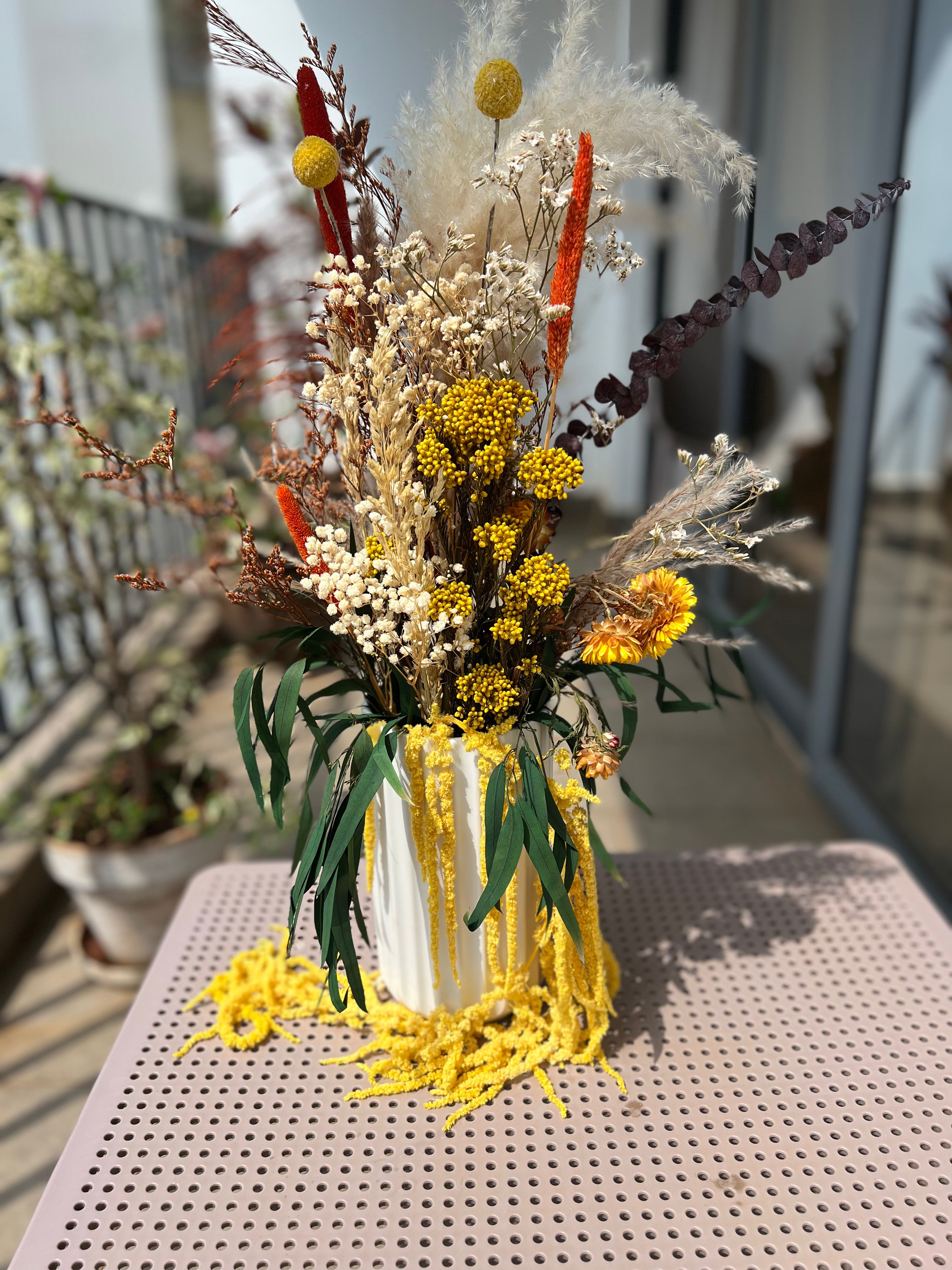 A decorative dried flower arrangement in a white ribbed vase, placed on a pink perforated metal table. The arrangement includes yellow mums, pampas grass, white baby's breath, orange stems, and green leaves, with yellow fringe hanging from the vase.
