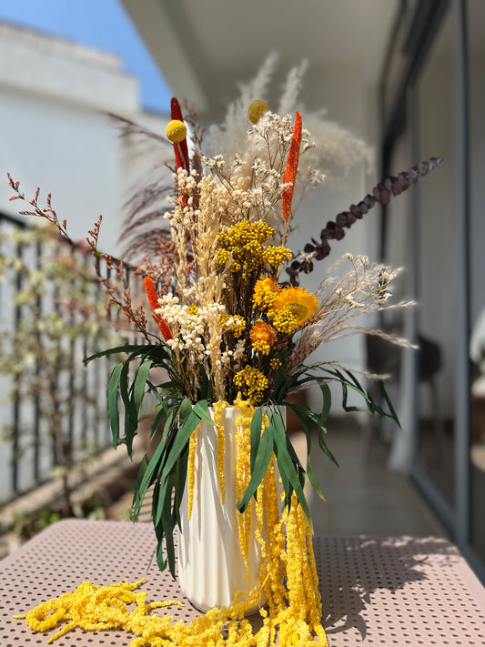 A dried flower arrangement from, kenyan lady fleur dried flowers kenya in a white ribbed vase, featuring yellow and orange flowers, beige pampas grass, and green leaves, displayed on a pink table outdoors.
