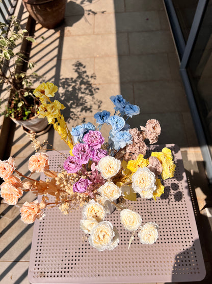 A colourful bouquet of real, preserved flowers from kenyan lady fleur flower shop of colours blue, beige, peach, white, yellow, and purple, in a white ribbed vase, placed on a light pink metal table in a sunlit balcony setting.
