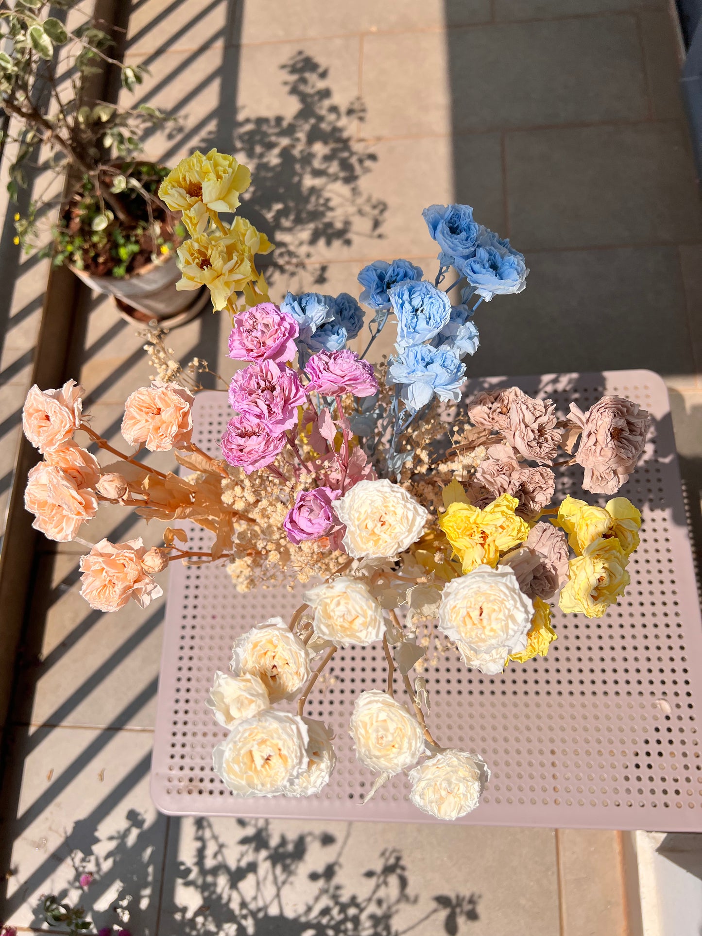 A colourful bouquet of real, preserved flowers of colours blue, beige, peach, white, yellow, and purple, in a white ribbed vase, placed on a light pink metal table in a sunlit balcony setting.