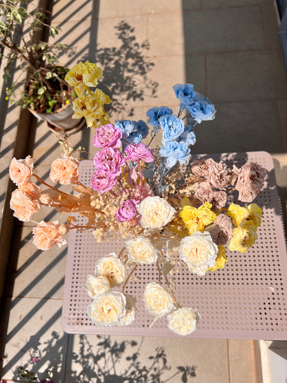 A colourful bouquet of real, preserved flowers of colours blue, beige, peach, white, yellow, and purple, in a white ribbed vase, placed on a light pink metal table in a sunlit balcony setting.