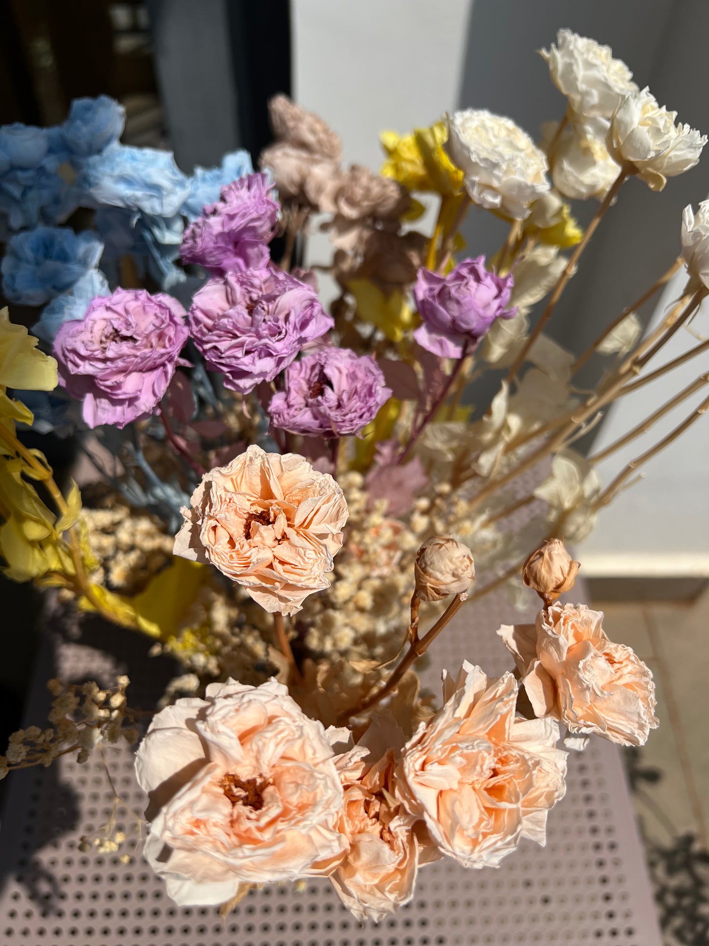 A bouquet of dried flowers in soft pastel tones, including peach roses, lavender blossoms, pale blue flowers, cream blooms, and yellow accents from kenyan lady fleur arranged on a perforated metal surface.