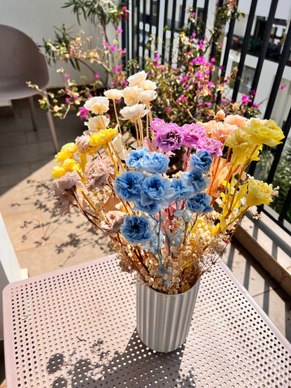 A colourful assortment of dried and preserved flowers in yellow, blue, pink, purple, and cream and short preserved baby's breath flowers in cream white from kenyan lady fleur arranged in a silver ribbed vase on a light pink perforated metal table outdoors.