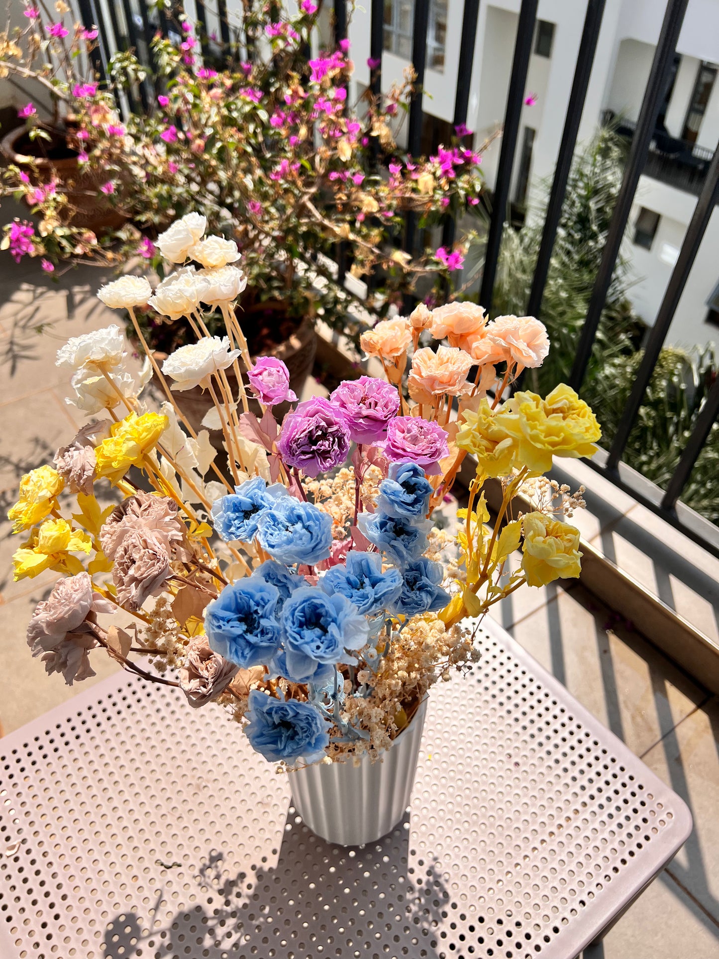 A colourful bouquet of real, preserved flowers of colours blue, beige, peach, white, yellow, and purple, in a white ribbed vase, placed on a light pink metal table in a sunlit balcony setting.