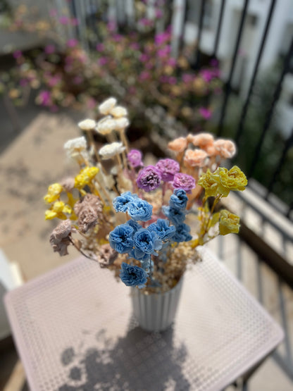 A small bouquet of dried and preserved  flowers from kenyan lady fleur in pastel colours including blue, purple, yellow, peach, cream, beige, and pink, arranged in a white ribbed vase on a light pink table with a blurred balcony background.