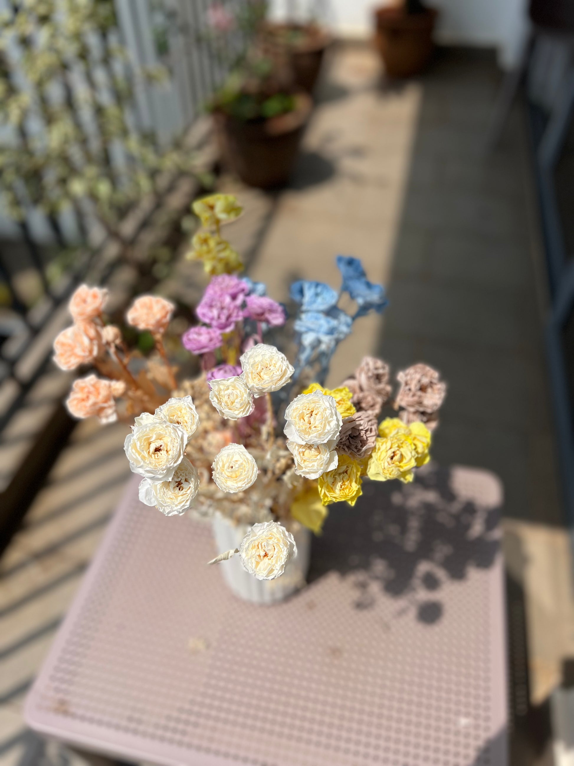 A small bouquet of dried and preserved flowers in pastel colours, including cream, yellow, blue, pink, peach, and lavender, arranged in a white ribbed vase on a pink table outdoors.