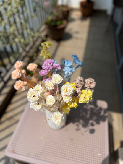 A small bouquet of dried and preserved flowers in pastel colours, including cream, yellow, blue, pink, peach, and lavender, arranged in a white ribbed vase on a pink table outdoors.