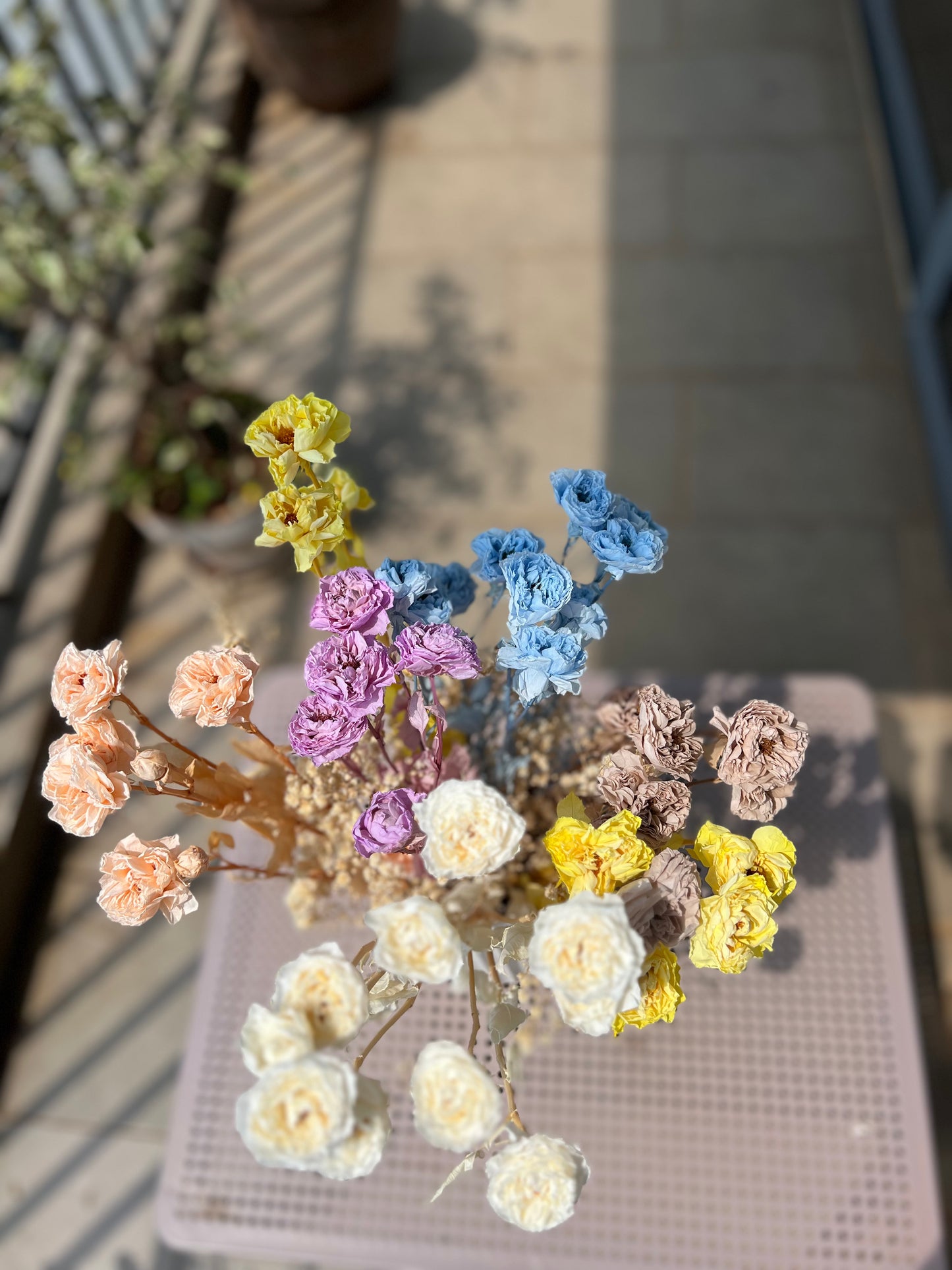A dried flower arrangement in a clear glass vase, featuring pastel-colored preserved wild roses blooms in blue, yellow, purple, peach, and cream, placed in an outdoor setting.