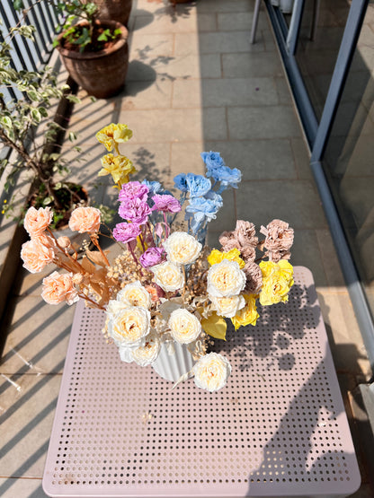 A colourful bouquet of real, preserved flowers of colours blue, beige, peach, white, yellow, and purple, in a white ribbed vase, placed on a light pink metal table in a sunlit balcony setting.