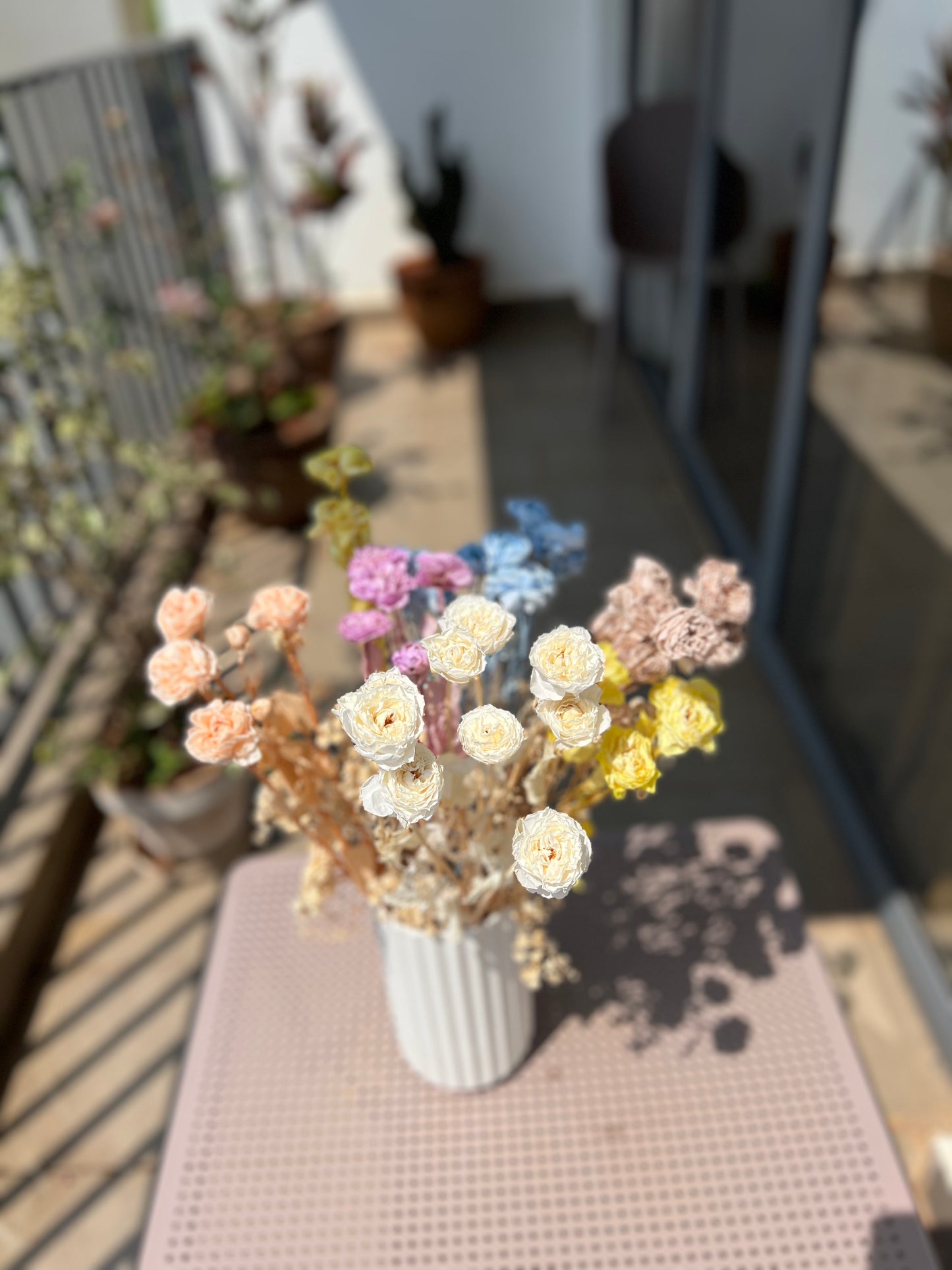 A bouquet of dried flowers from kenyan lady fleur in pastel shades of cream, pink, purple, blue, and yellow arranged in a white ribbed vase, placed on a pink textured mat on a balcony with a blurred outdoor background.