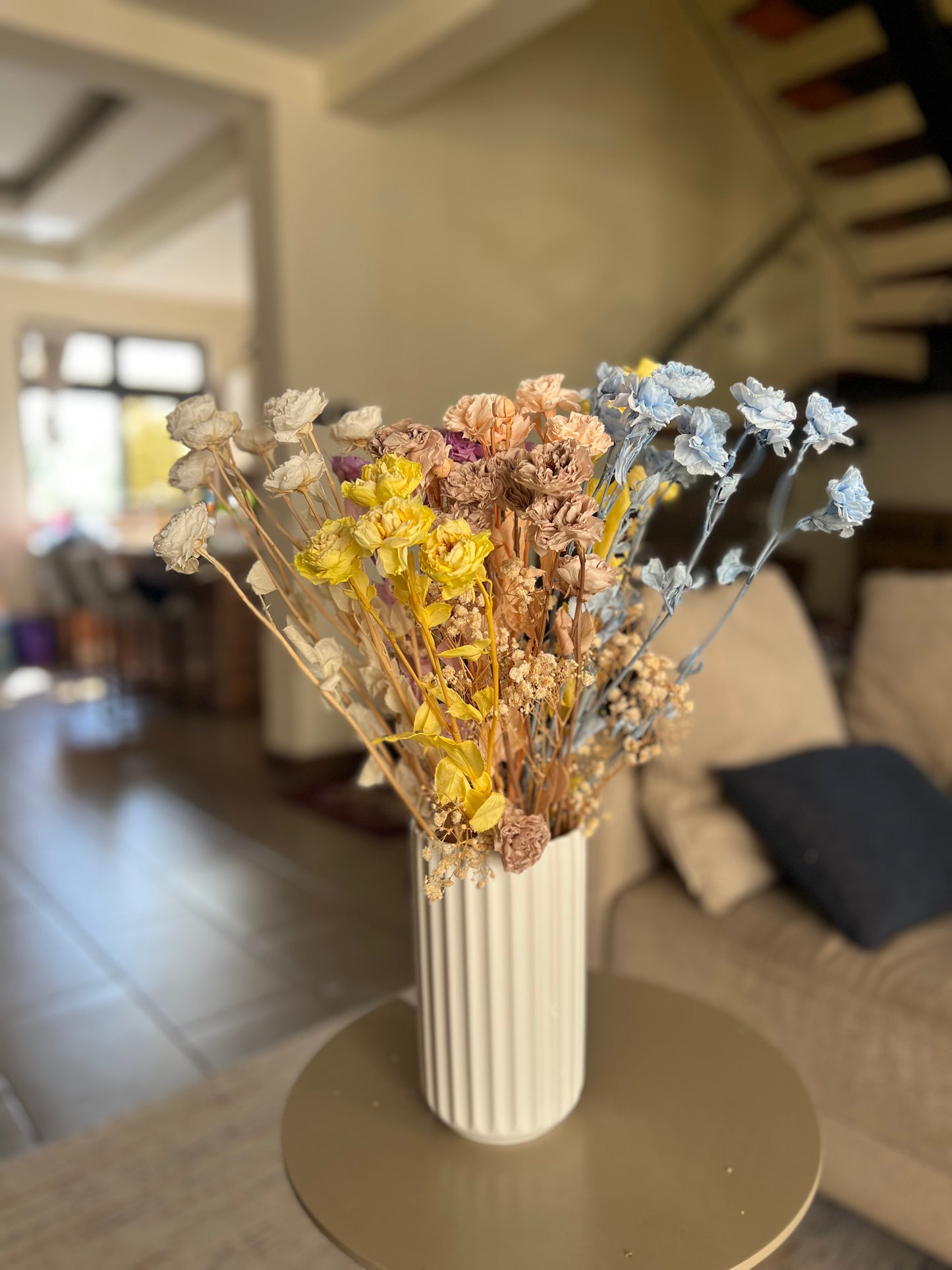 A white ribbed ceramic vase filled with a dried flower arrangement in soft pastel tones from kenyan lady fleur dried flowers kenya placed on a round table in a blurred living room setting.