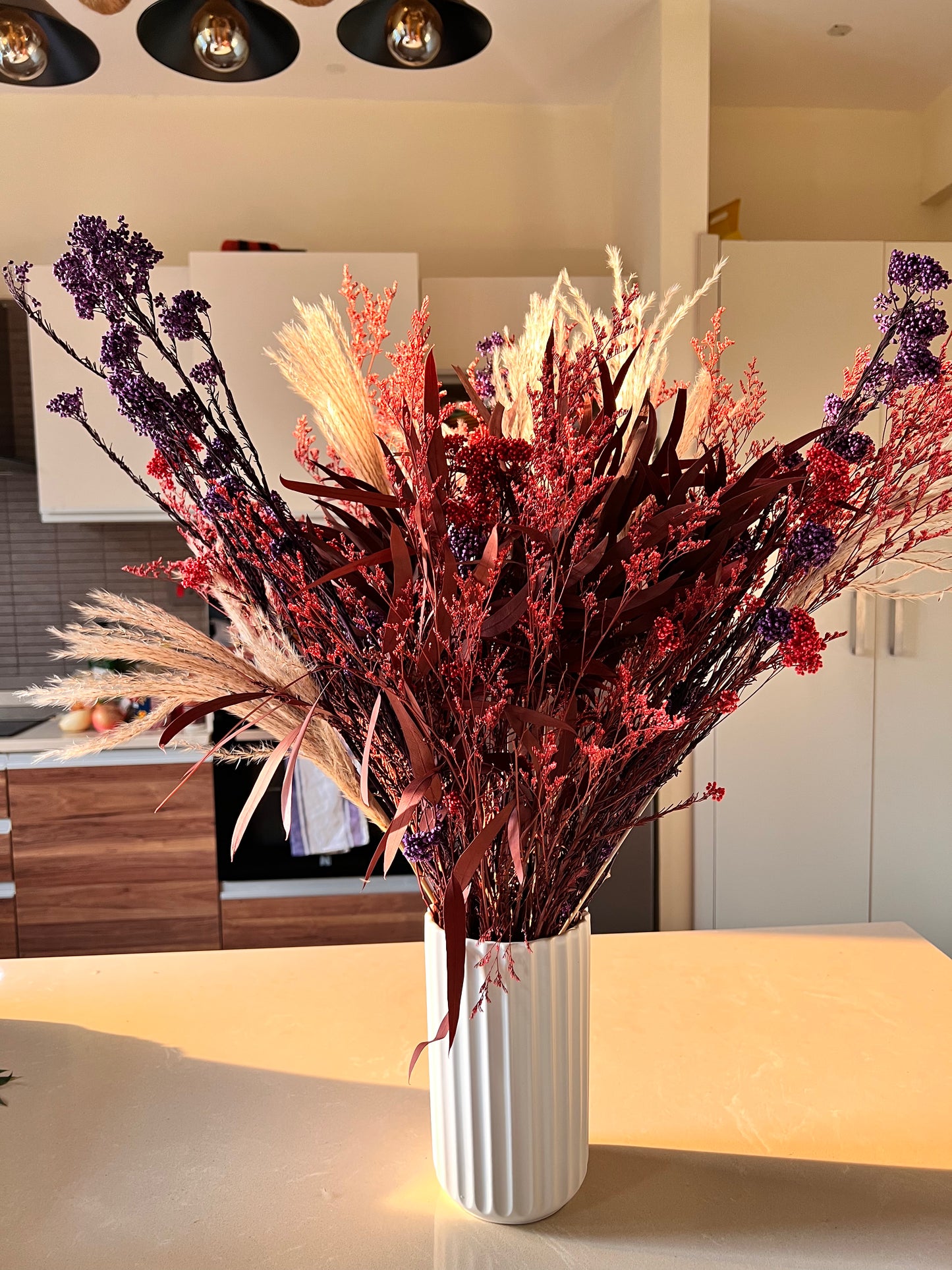 A dried floral arrangement in a white ribbed ceramic vase, featuring red berries, purple sprigs, and beige pampas grass, displayed on a kitchen island.