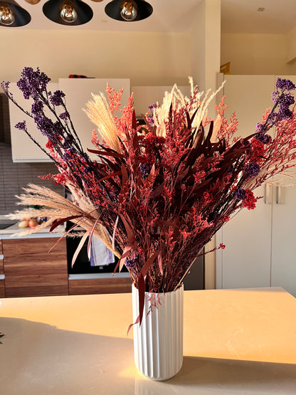 A dried floral arrangement in a white ribbed ceramic vase, featuring red berries, purple sprigs, and beige pampas grass, displayed on a kitchen island.