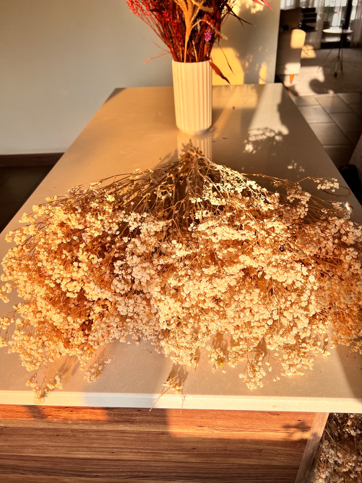 A large arrangement of preserved dried baby's breath flowers in a warm beige and brown tone from kenyan lady fleur dried flowers kenya, displayed on a light-colored wooden table in a home interior setting with a white ribbed vase in the background.