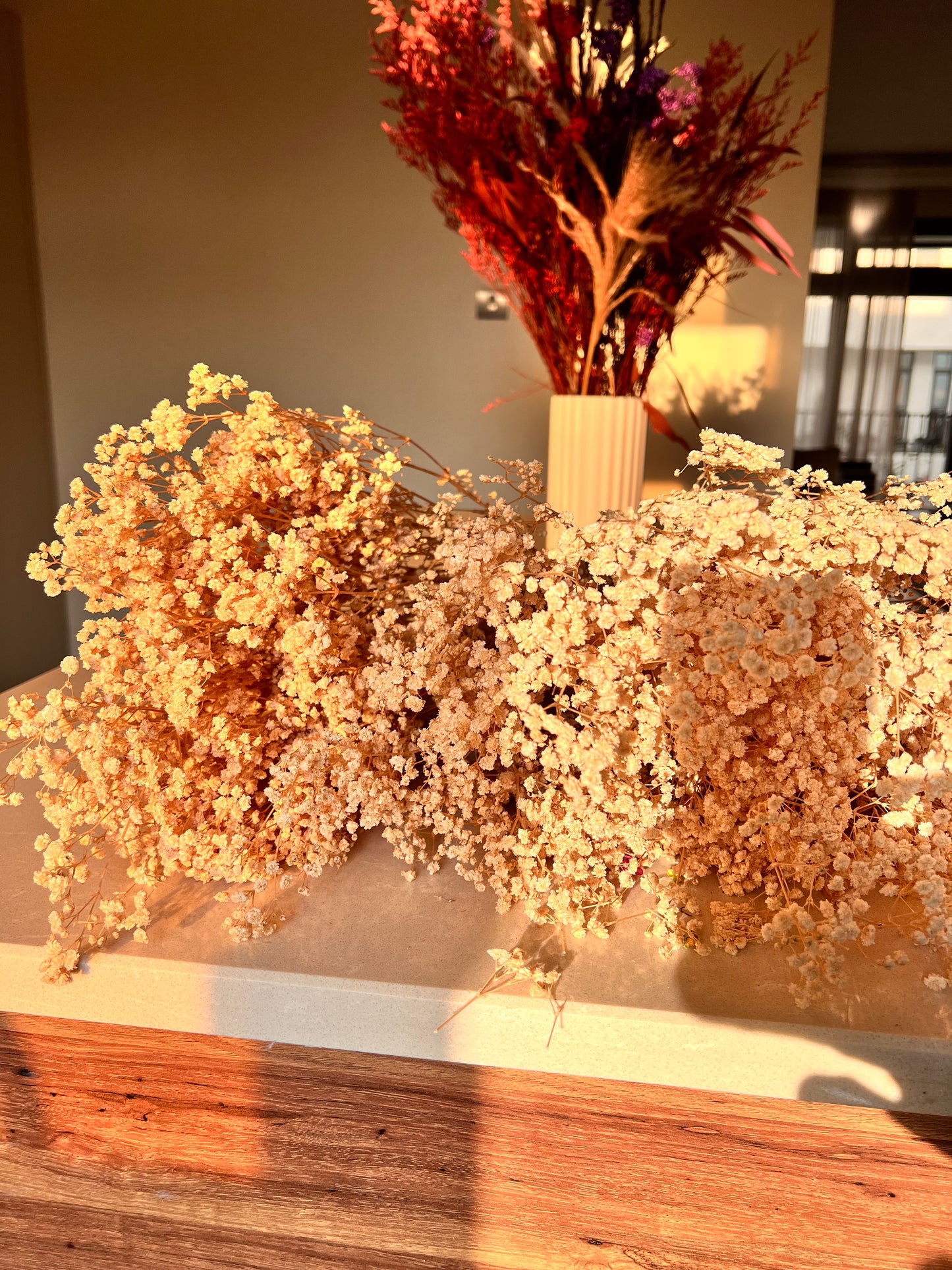 Dried floral arrangement featuring a large cascading cluster of cream preserved baby's breath from kenyan lady fleur of dried flowers kenya in the foreground and a smaller vase with red and purple dried stems in the background, set on a wooden surface.