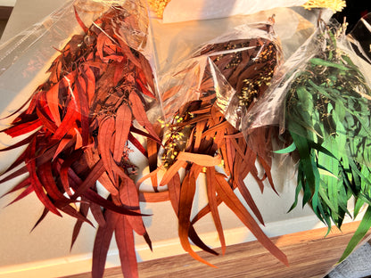 Three bundles of dried decorative botanicals wrapped in clear plastic, showing red, brown, and green dried leaves and seed pods on a light surface.
