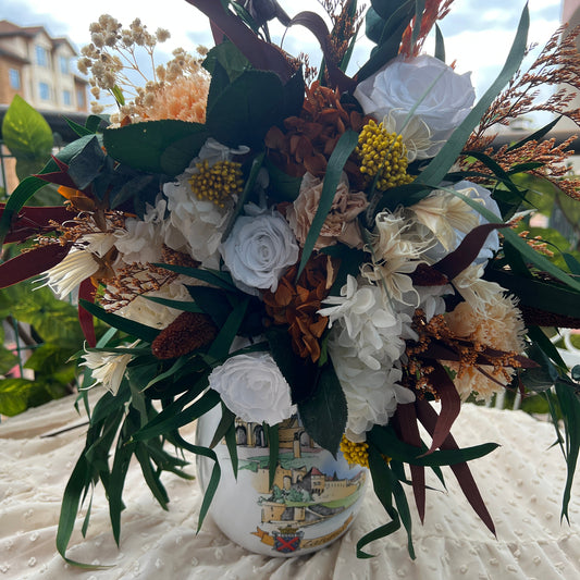 A decorative floral arrangement in a white ceramic vase with a printed landscape design, placed on a textured cream tablecloth outdoors with buildings and sky blurred in the background.