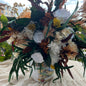 A decorative floral arrangement in a white ceramic vase with a printed landscape design, placed on a textured cream tablecloth outdoors with buildings and sky blurred in the background.