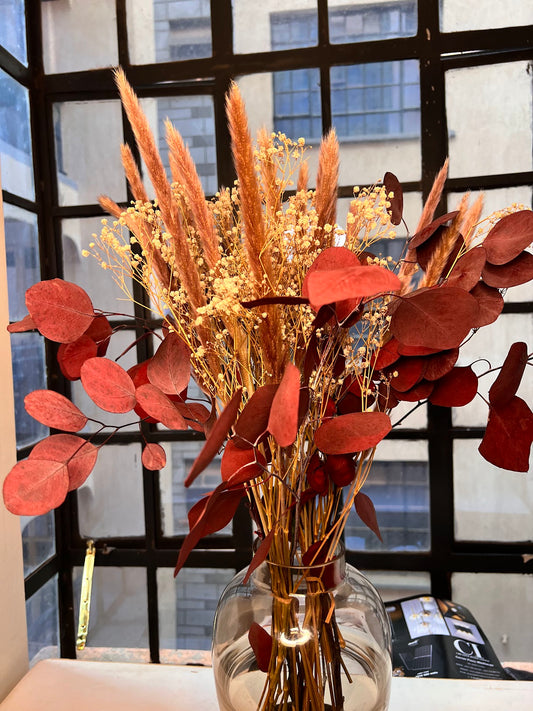 Decorative bouquet with red leaves and gold stems in a clear vase against a window background.
