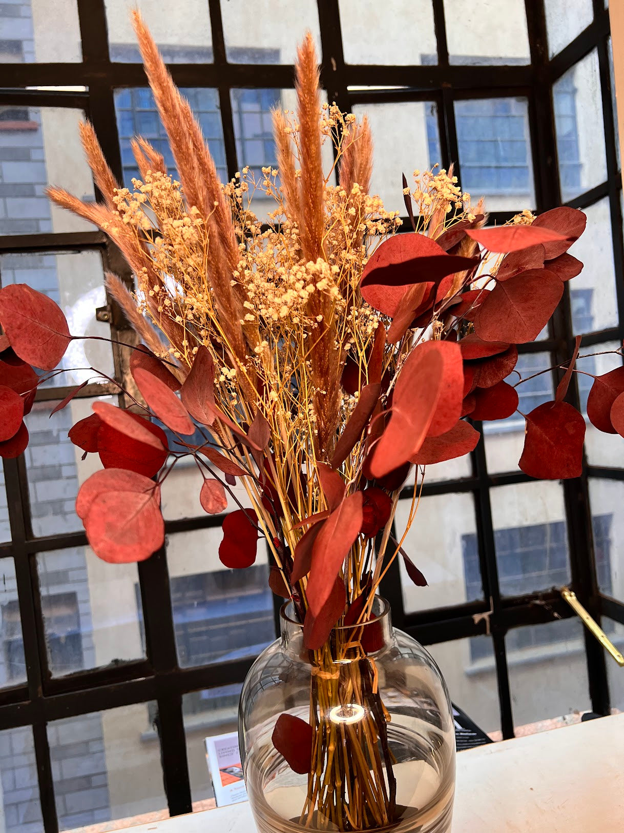 Clear vase with dried flowers and brick red eucalyptus leaves in front of a window with a cityscape view.