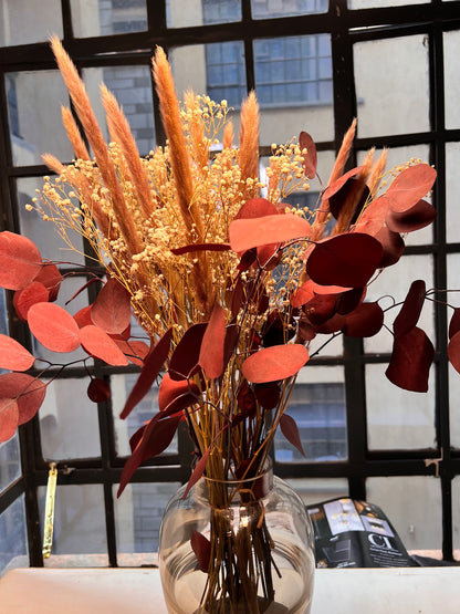 Decorative bouquet with red leaves and brown hue fillers in a clear vase against a windowed from kenyan lady fleur.
