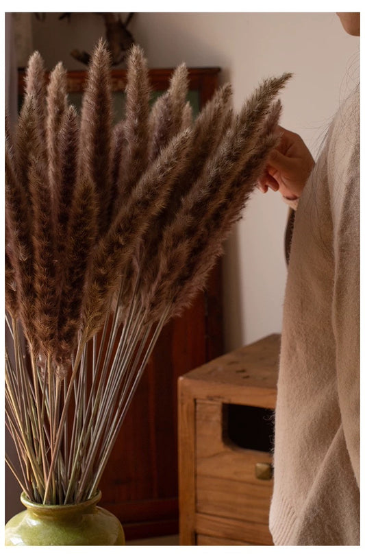 Person holding a bundle of dried natural pampas grasses in a vase in front of a wooden cabinet.