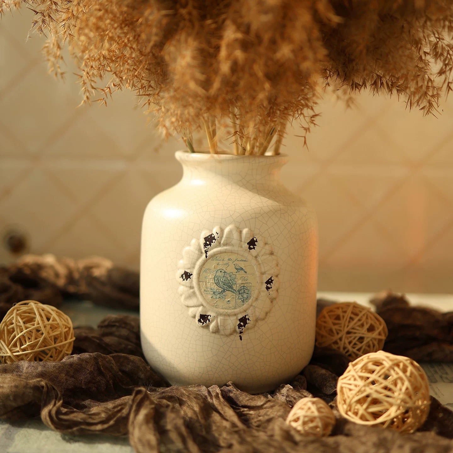 Decorative white vase with a decorative emblem surrounded by dried plants and spherical decorations on a textured surface.
