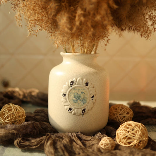 Decorative white vase with a decorative emblem surrounded by dried plants and spherical decorations on a textured surface.