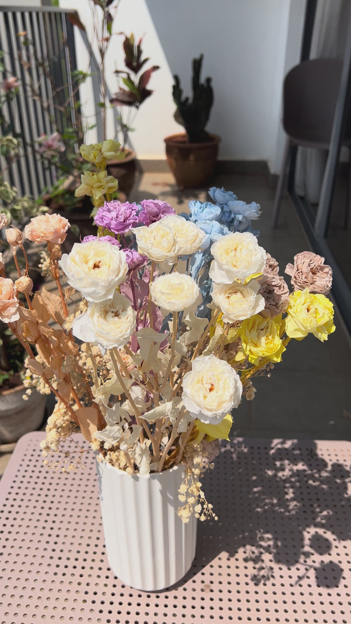 video of a colourful bouquet of real, preserved flowers of colours blue, beige, peach, white, yellow, and purple, in a white ribbed vase, placed on a light pink metal table in a sunlit balcony setting.