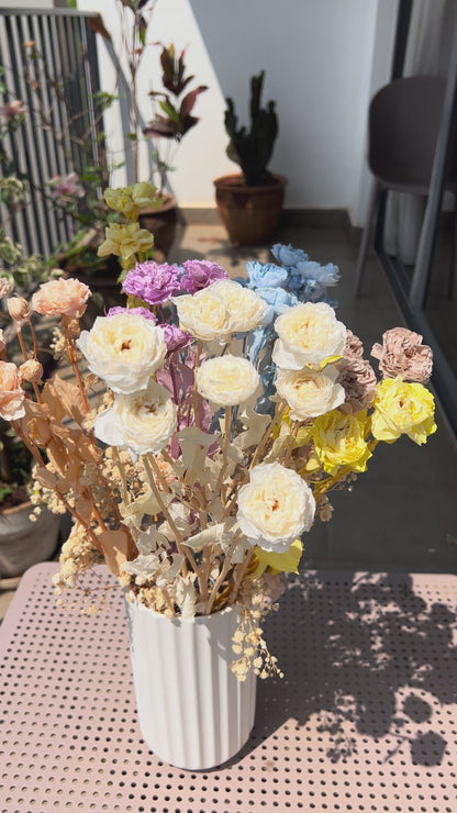 video of a colourful bouquet of real, preserved flowers of colours blue, beige, peach, white, yellow, and purple, in a white ribbed vase, placed on a light pink metal table in a sunlit balcony setting.