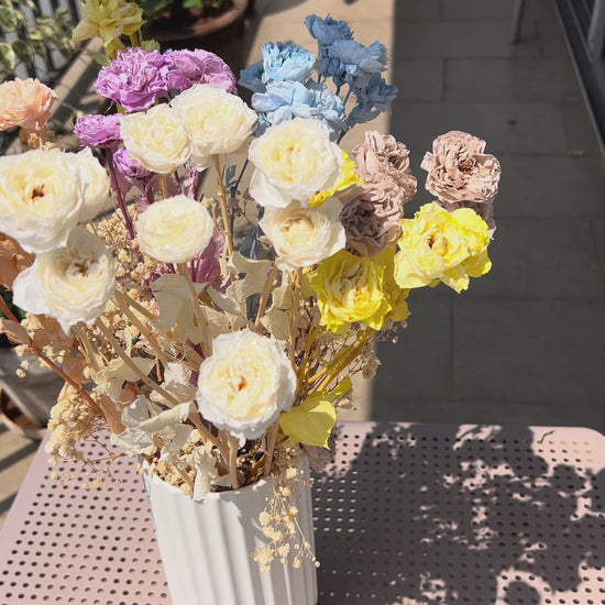 video showing a colourful bouquet of real, preserved flowers from kenyan lady fleur flower shop of colours blue, beige, peach, white, yellow, and purple, in a white ribbed vase, placed on a light pink metal table in a sunlit balcony setting.
