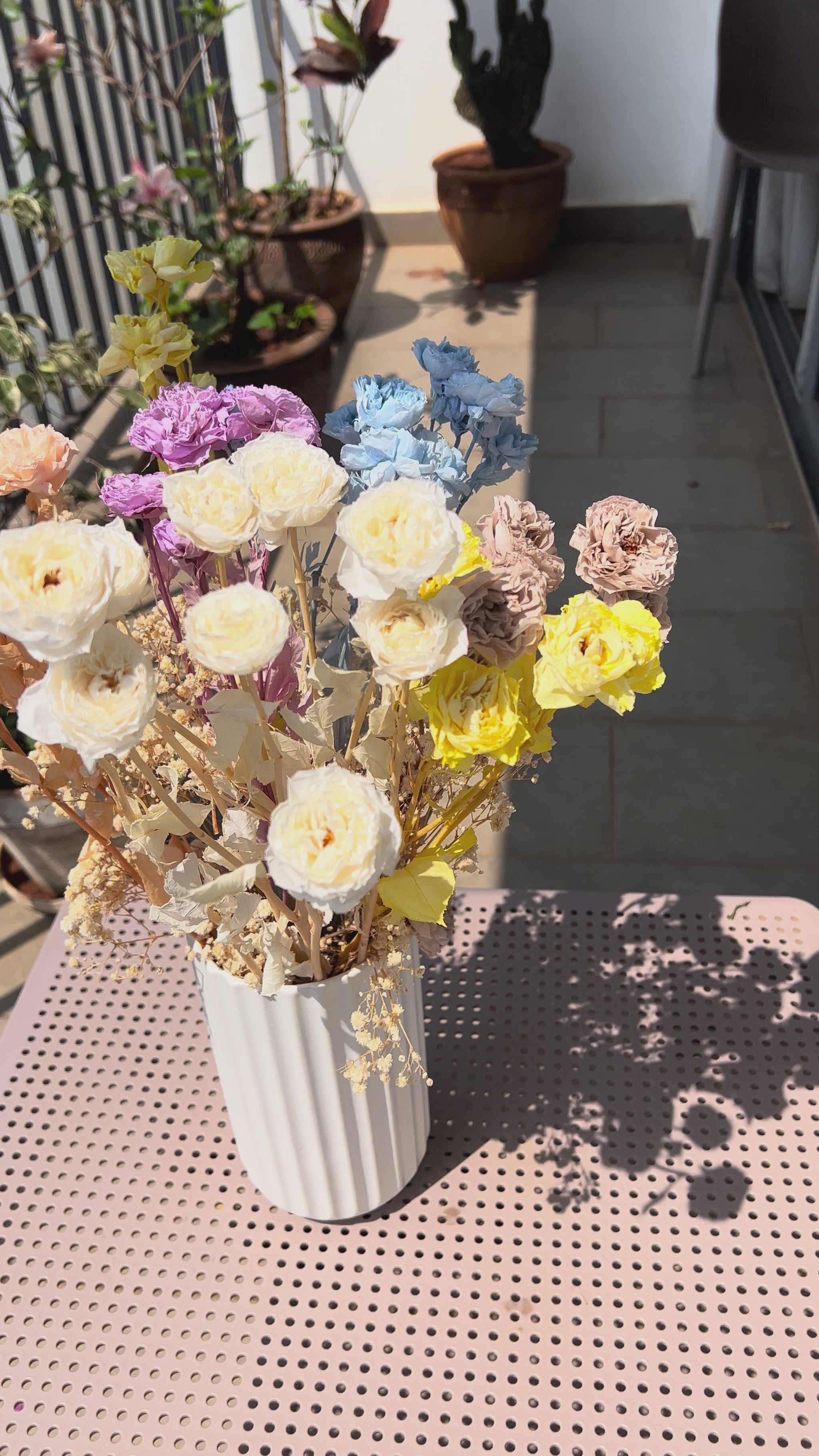 video showing a colourful bouquet of real, preserved flowers from kenyan lady fleur flower shop of colours blue, beige, peach, white, yellow, and purple, in a white ribbed vase, placed on a light pink metal table in a sunlit balcony setting.