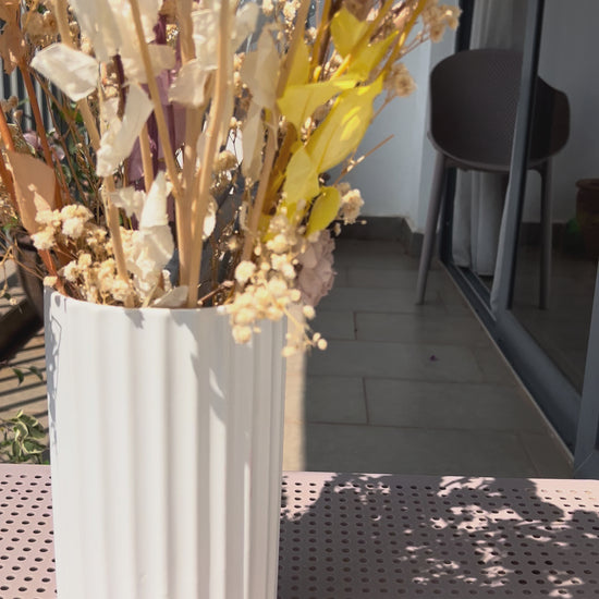 video of a colourful bouquet of real, preserved flowers of colours blue, beige, peach, white, yellow, and purple, in a white ribbed vase, placed on a light pink metal table in a sunlit balcony setting.