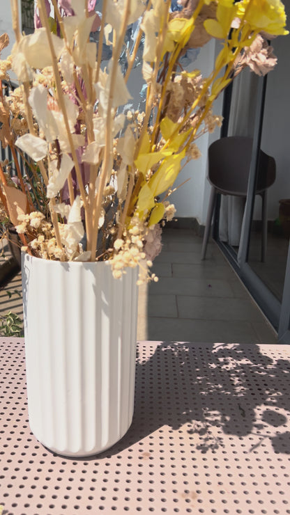 video of a colourful bouquet of real, preserved flowers of colours blue, beige, peach, white, yellow, and purple, in a white ribbed vase, placed on a light pink metal table in a sunlit balcony setting.
