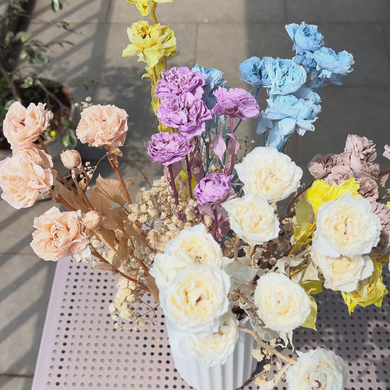 A video of a colourful bouquet of real, preserved flowers of colours blue, beige, peach, white, yellow, and purple, in a white ribbed vase, placed on a light pink metal table in a sunlit balcony setting.