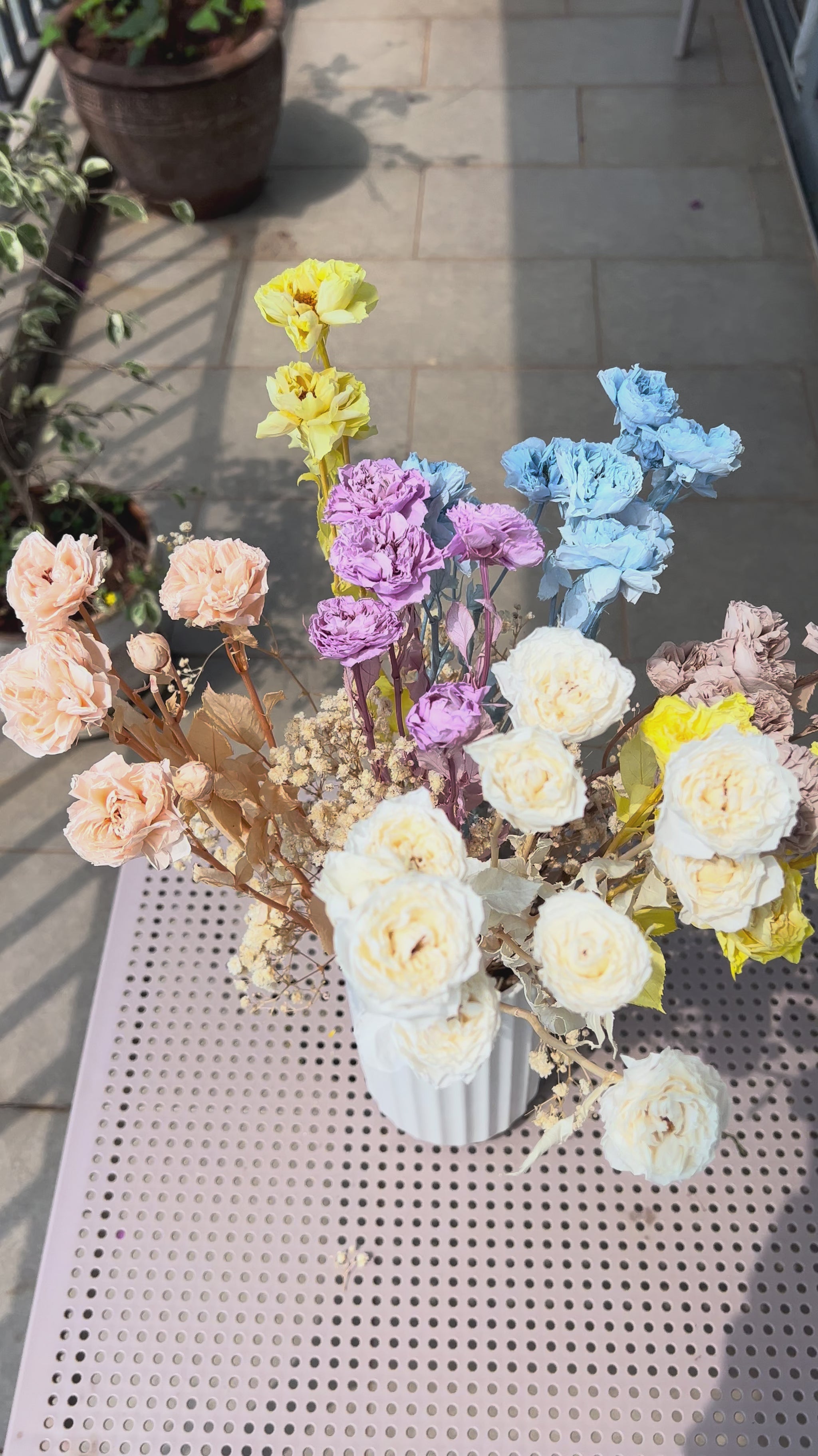 A video of a colourful bouquet of real, preserved flowers of colours blue, beige, peach, white, yellow, and purple, in a white ribbed vase, placed on a light pink metal table in a sunlit balcony setting.